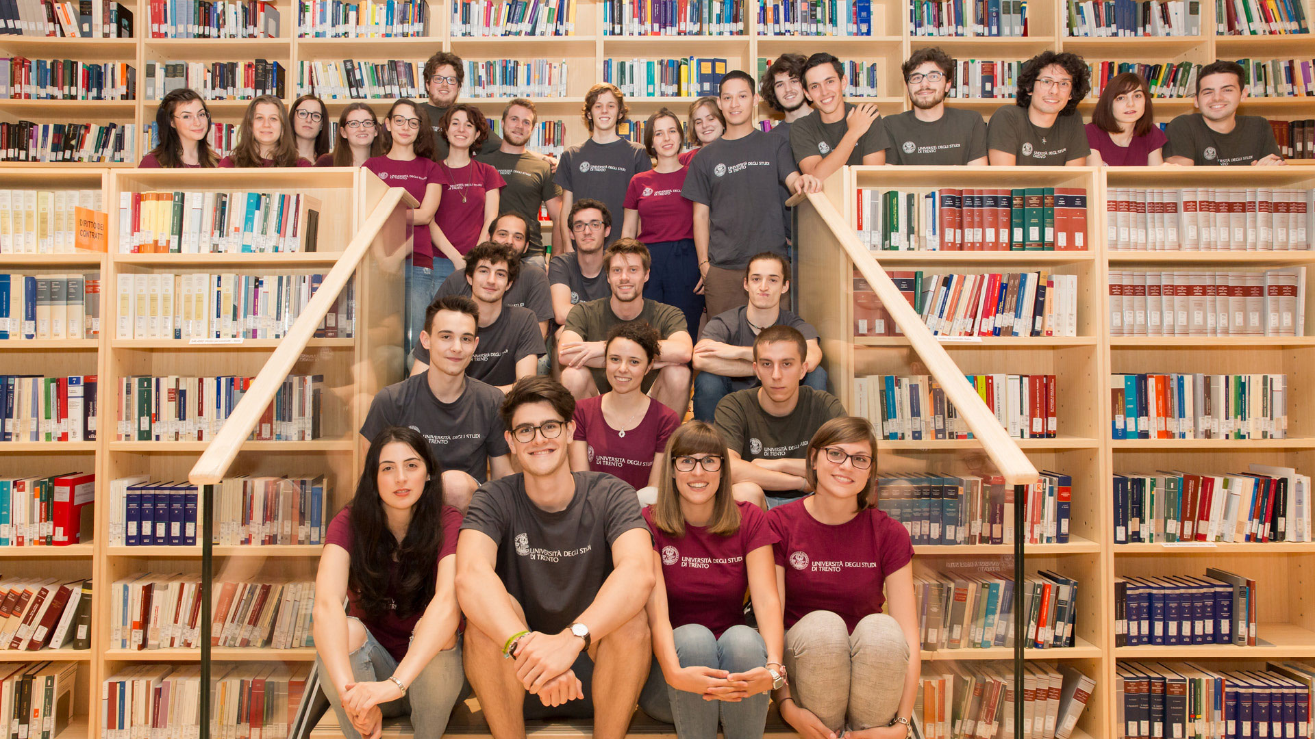 Gli orientatori di UniTrento: un gruppo di ragazzi e ragazze, seduti sulla scalinata della biblioteca. Indossano la t-shirt bordeaux di UniTrento Orienta e sorridono