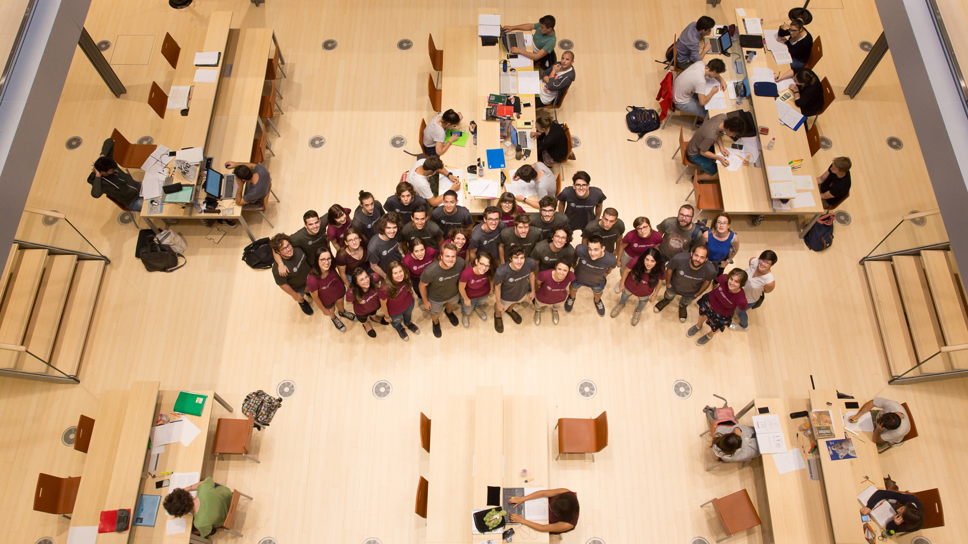 Un gruppo di studenti e studentesse orientatori UniTrento ripresi dall'alto di una scala, all'interno della Biblioteca. Guardano in alto, intorno a loro tavoli con studenti che studiano e leggono.