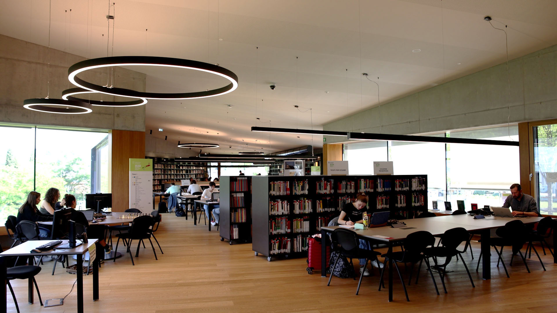 L'interno della Biblioteca universitaria di Mesiano, a scaffale aperto, con tavoli per lo stdio individuale e di gruppo e gradi vetrate con vista sul parco e la città di Trento