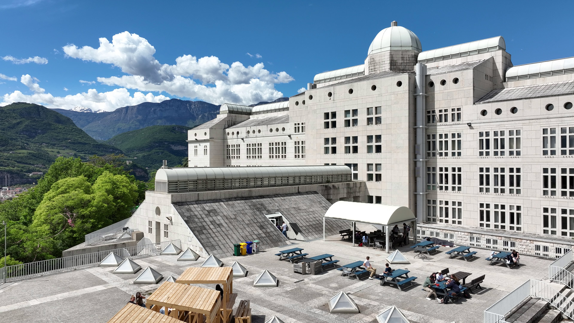 L'edificio principale del Dipartimento di Ingegneria Civile Ambientale e Meccanica: un grande edificio bianco all'interno di un parco. A sinistra e sullo sfondo, la vegetazione e le montagne verdi, il cielo azzurro 