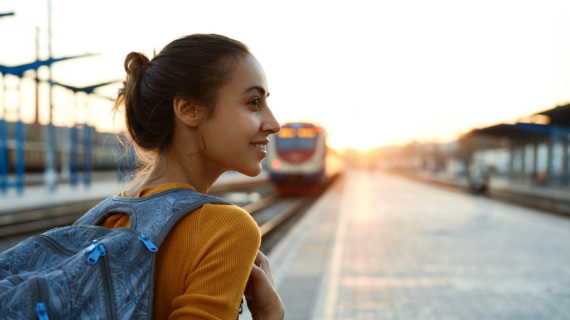 Una ragazza con lo zaino sulle spalle, girata a 3/4, va verso un treno in arrivo sullo sfondo